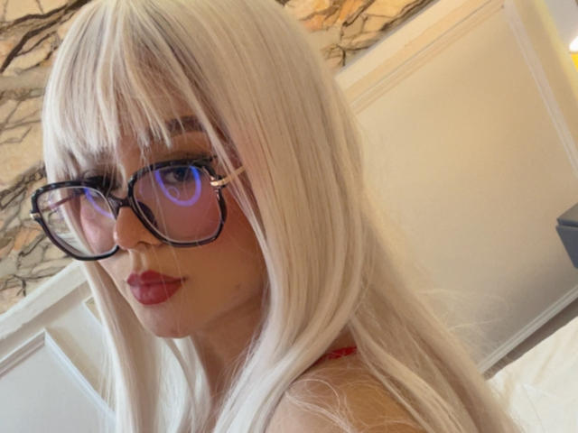 Millie Reed, con su largo cabello blanco, gafas y lápiz labial rojo, mostrando su linda cara y mirando por encima del hombro. Sus glúteos no son visibles..