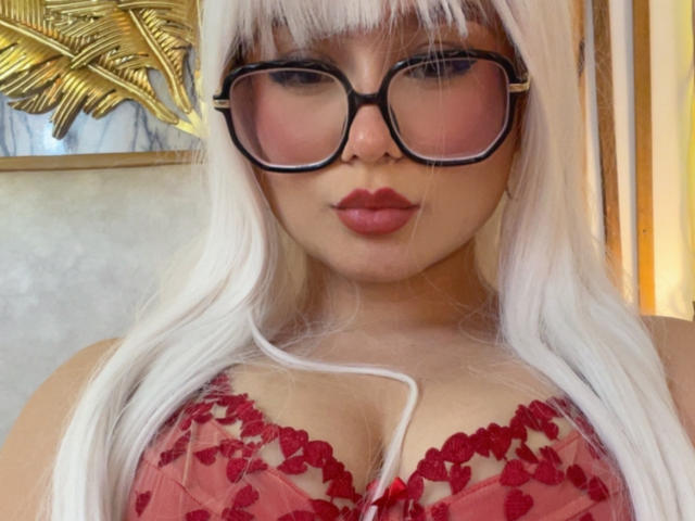 Millie Reed, avec ses longs cheveux blancs, ses lunettes noires et son soutien-gorge en dentelle rouge, exhibe ses seins gros, brillants et aux mamelons rouges. Ses lèvres sont pulpeuses et elle regarde l'appareil photo..