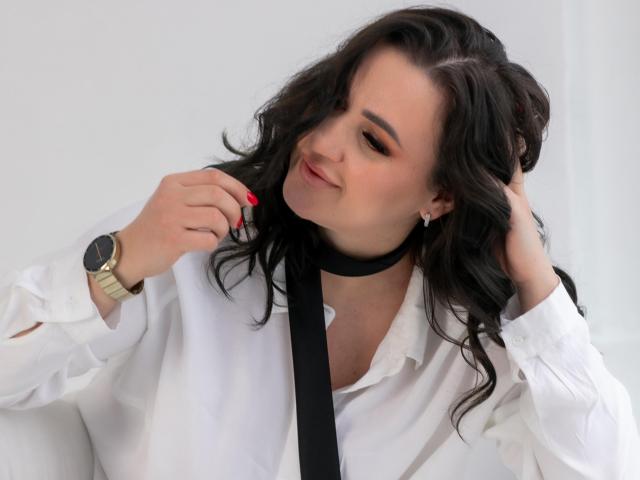 AnnabelleAllie with wavy black hair, white shirt, black tie, gold watch, red nail polish, and a slight smile, adjusting her tie with one hand.