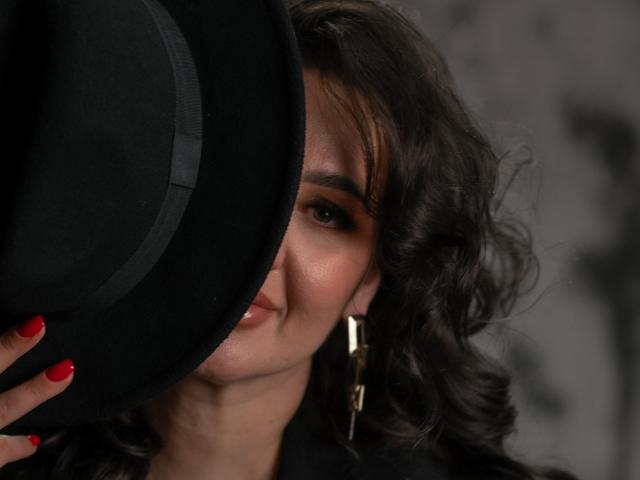 AnnabelleAllie holding a black hat, partially covering her face, with red nails, curly dark hair, and gold earrings, showing a subtle smile. Her ass is not visible.