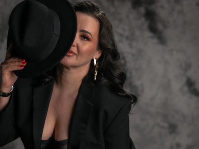 AnnabelleAllie with wavy black hair, holding a black hat over her face, wearing a black blazer and showing cleavage, gold earrings, red nails, and a bracelet.