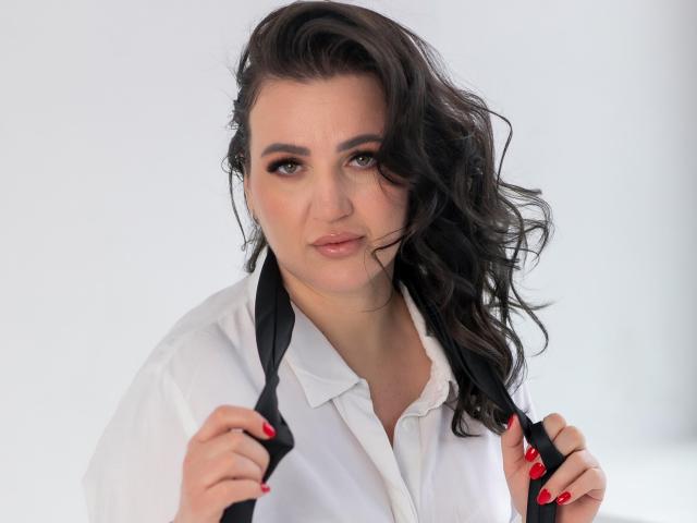 AnnabelleAllie with wavy black hair, white shirt, holding black tie, red nails, slight smile, looking at camera, background blurred. Her tits are medium-sized, she's cute and sexy.