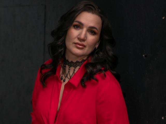 AnnabelleAllie with curly black hair, wearing a red shirt, black lace choker, and subtle makeup, looking slightly to the side with a gentle smile. Boobs visible under shirt.