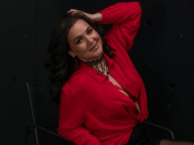 AnnabelleAllie with long black hair, wearing a red button-up shirt, black lace choker, and earrings, smiling, hand in hair, sitting on a chair, showing a bit of cleavage.