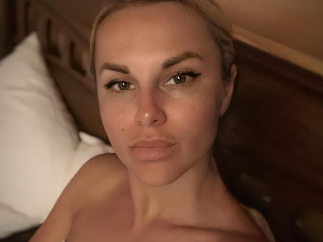 BelleLisaG heeft een serieus gezicht met bruine ogen, zwarte eyeliner en lichte huid, en kijkt omhoog naar de camera, ongetooid, met een wit kussen op de achtergrond..
