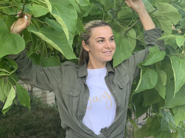 BelleLisaG with green leaves in her hands, smiling, wearing an open green shirt over a white tee with "joy" in gold. She has blonde hair and green eyes.