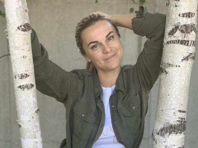 BelleLisaG with arms raised, holding white birch trees, wearing an olive green jacket, and a white shirt, smiling with green eyes, and brown hair in a ponytail.