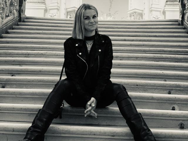 BelleLisaG with straight blonde hair, black leather jacket, and boots, sitting on stone stairs, legs spread, hands between knees, looking at camera, black and white photo.