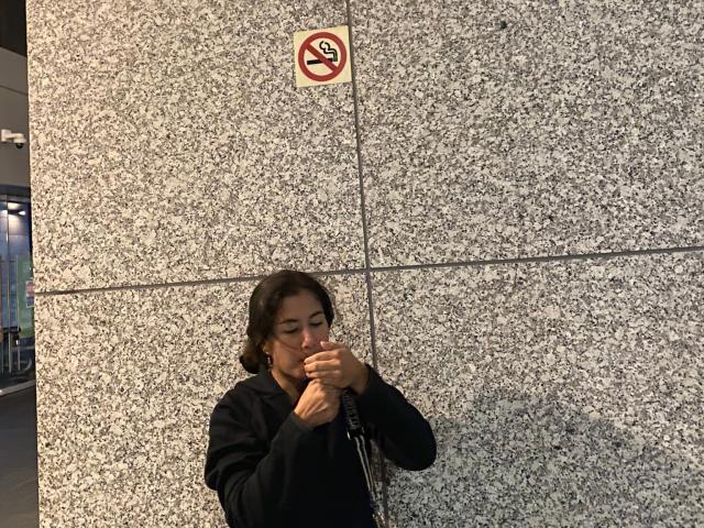 AuroraMonroe, with medium-length brown hair, wearing a black hoodie, smoking a cigarette, standing against a speckled gray wall, looking downward with a serious expression.