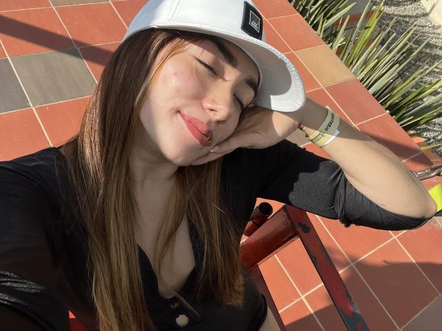 SharonKlart with eyes closed, wearing a white cap, black top, and pink lipstick, resting her head on her hand, showing a slight smile and her small tits.
