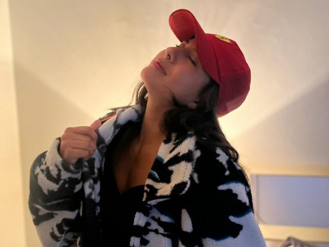AuroraMonroe with her head tilted back, wearing a red cap, black and white camo jacket, and black top, her nose pierced, and brown wavy hair falling.