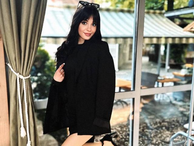 MySoftLinessa, con el pelo largo y negro, vestida con un traje negro, sosteniendo una cortina, de pie frente a una ventana, con tacones negros y gafas de sol en la cabeza..