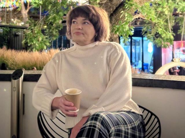 NelyGratitu with short brown hair, wearing a white sweater and black plaid skirt, holding a coffee cup, sitting outside, looking to the side with a slight smile.