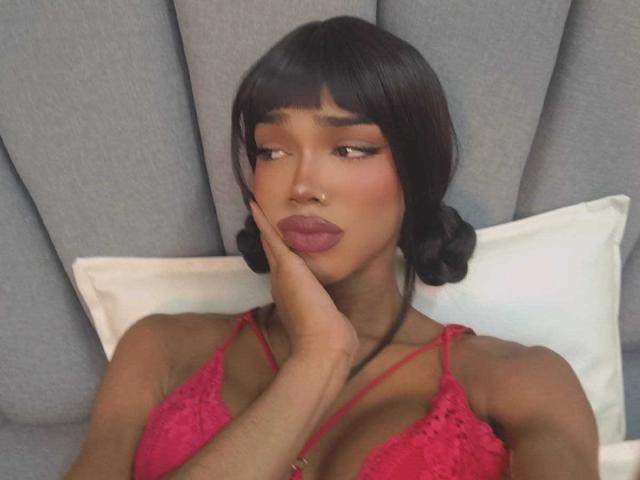 Naomi Steward mit dunkelbraunen Zöpfen, trägt einen roten Spitzen-BH, ihre Hand berührt ihre Wange, sie blickt zur Seite mit rosafarbenen Lippen. Brüste sind sichtbar..