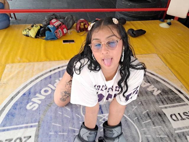 EmiilyJohamson, tongue out, wearing glasses, white shirt, knee pads, black boots, and dog ears, has tattoos on her arm, in a wrestling ring, with sports gear around.
