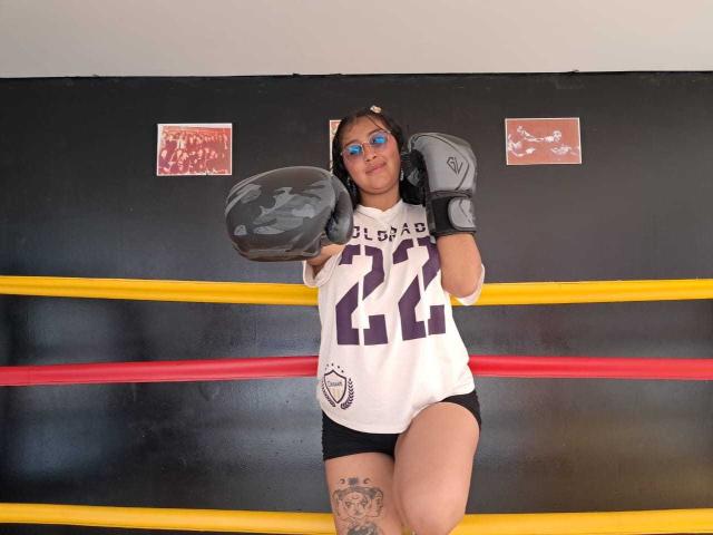 EmiilyJohamson with black braided hair, wearing a white jersey with "BLB" and "44", black shorts, and gray boxing gloves, standing in a boxing ring, showing a tattoo on thigh.