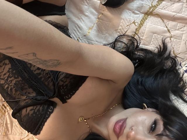 ThaliaCollins con lingerie in pizzo nero, che mostra il seno, sdraiata su un letto trapuntato, capelli scuri, collana d'oro, tatuaggio sul braccio e un'espressione sexy e seducente..