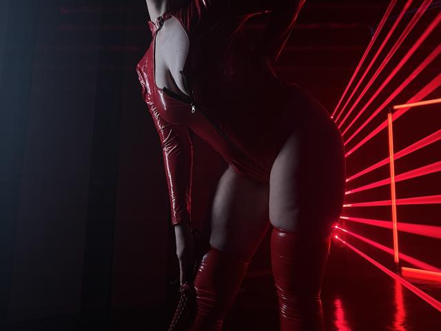 GoddessSophie69 in a shiny red latex bodysuit, exposing her tits with a deep cleavage, and red thigh-high boots, leaning forward with her ass up, under red neon lights.