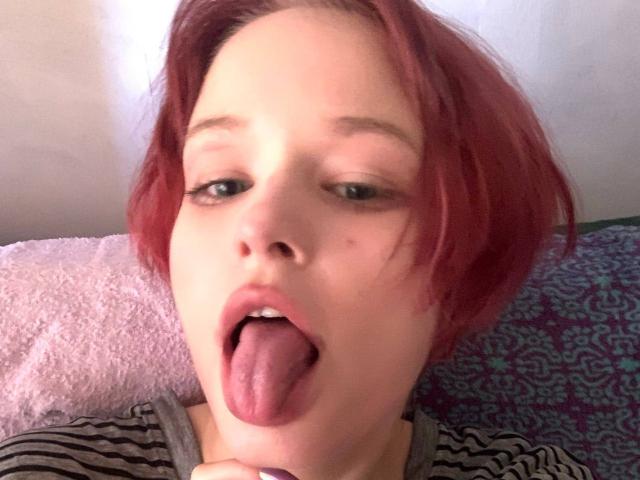 Close-up photo of ElisKeller with short, red-dyed hair, light skin, and small, slightly upturned nose. ElisKeller's tongue is extended, sticking out slightly. She has thin, arched eyebrows and light brown eyes looking slightly to the left. She's wearing a striped shirt, and her right hand is near her chin. The background includes a pink textured pillow and a blue patterned fabric. The lighting is soft, highlighting her facial features.