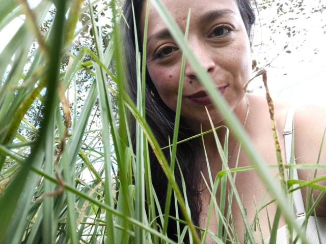 StangelarRivera's face peeks through tall green grass with a subtle smile, brown eyes, and long dark hair, wearing a white tank top, her small tits barely visible.
