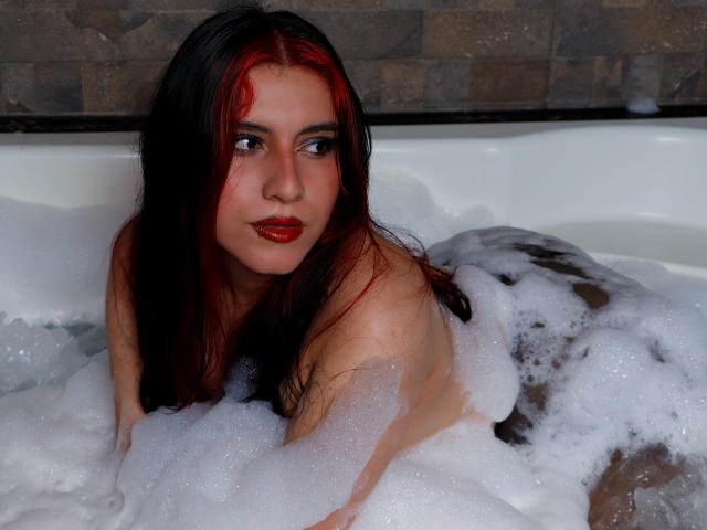 DannaOlivieri with red and black hair, red lips, and soap bubbles in a bathtub, lying on her stomach, showing her bare back and buttocks. Her eyes look forward.