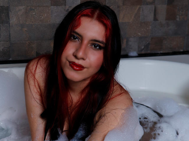 DannaOlivieri with red and black hair, sitting in a bubble-filled bathtub, smiling with red lipstick, her bare shoulders visible, looking directly at the camera.