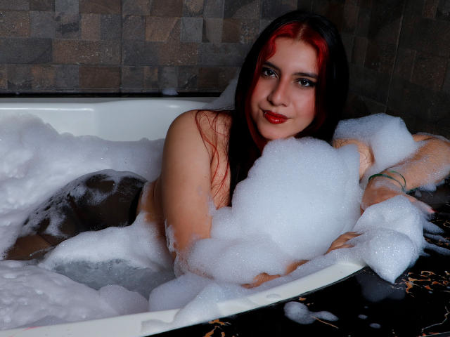 DannaOlivieri with red and black hair, naked, in a bubble-filled bathtub, smiling at the camera, showing her tits and arms, with foam covering her ass.