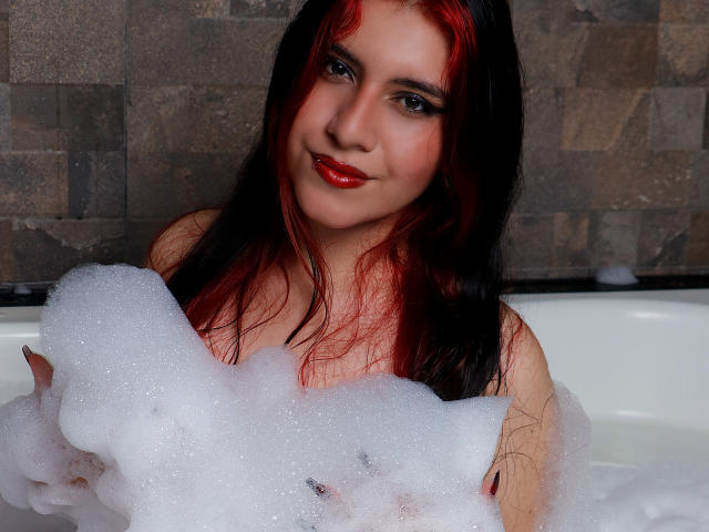DannaOlivieri with red-black hair, red lipstick, and bubble-filled bathtub, smiling, holding foam over her tits, visible in the white tub.