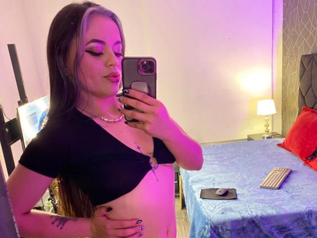 ZoeyStone with purple highlights, black crop top, and pearl necklace, showing her small tits in a mirror selfie, holding a phone, in a bedroom with blue bed and lamp.