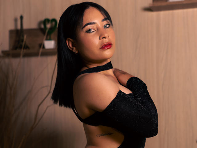 SaharaArisa with black bob, red lipstick, and sparkly black top, covering her tits with arms, tattoo on right side, wooden background, looking at camera with sultry expression.