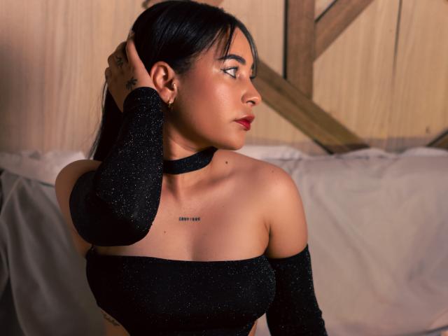 SaharaArisa with dark hair, wearing a sparkly black off-shoulder top, looking to the side, hand in hair, showing her tits and small black tattoo on chest.