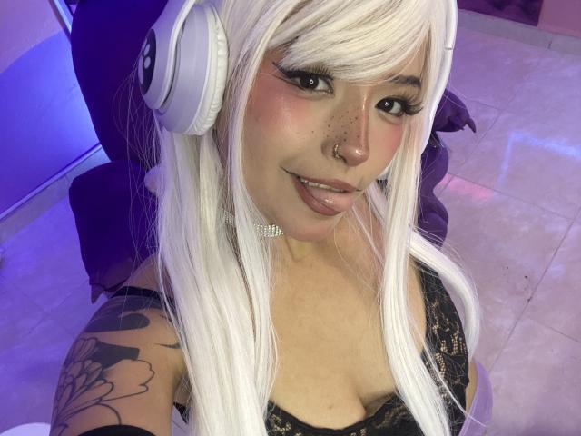 CarlaMiceli with white hair, headphones, black lace top, tattoo on left arm, nose ring, and tongue out, has pink blush and freckles, showing her tits.