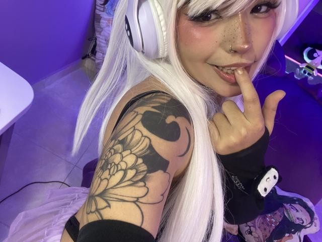CarlaMiceli with long white hair, nose ring, and finger in mouth, showing a tattooed arm, wearing headphones, and black fingerless glove, in a purple-lit room.
