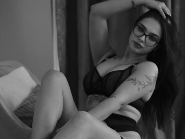 SienaFoxxy in black lingerie, glasses, and a tattoo on her arm, sitting with legs up, hand in hair, looking at camera, showing her tits and a cute ass.