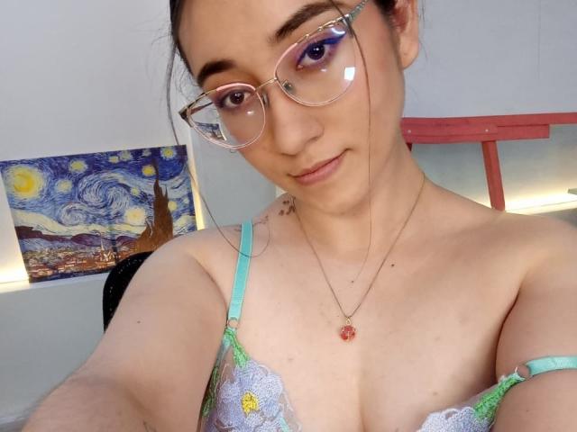 SwettieSammy with glasses, light skin, and a subtle smile, wearing a green and purple lace bra, showing her tits. Painting of "Starry Night" in the background.