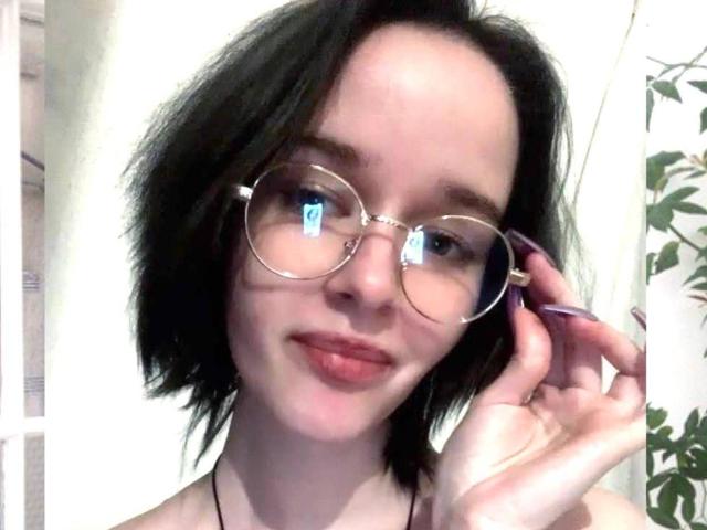 Elis Keller tem cabelo preto curto, usa óculos redondos e está com a língua para fora, com uma mão perto do rosto. Ela tem pele clara e está usando um colar preto..