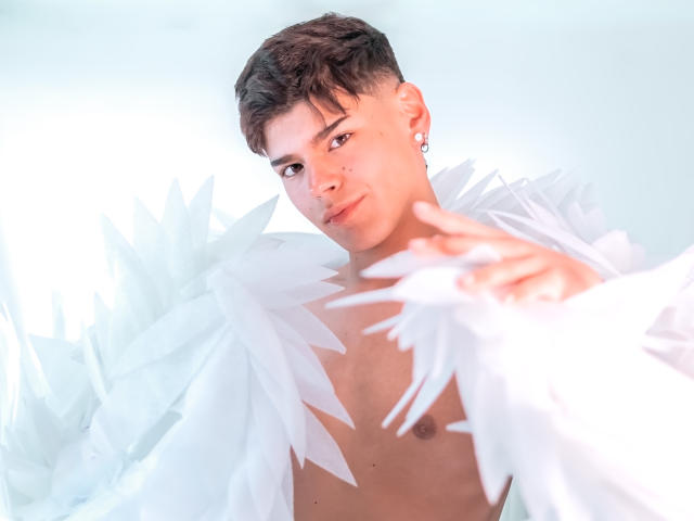 KevinMiet with short brown hair, wearing white feathered garment, shirtless, small chest, ear piercings, reaching forward, soft light, slight smile, focused expression, white background.