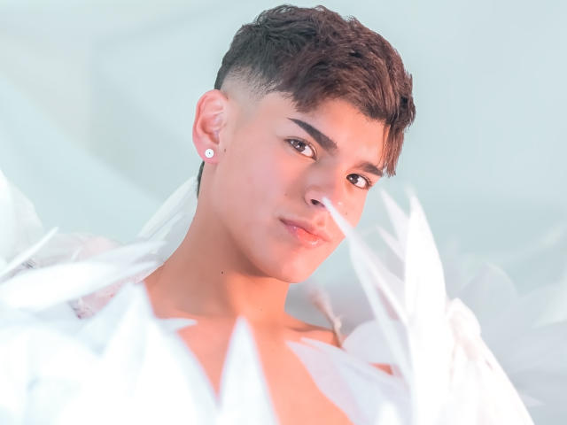 KevinMiet with short brown hair, light skin, and a serious expression, wearing white feathers, has a small white earring in his right ear. Soft light highlights his face.