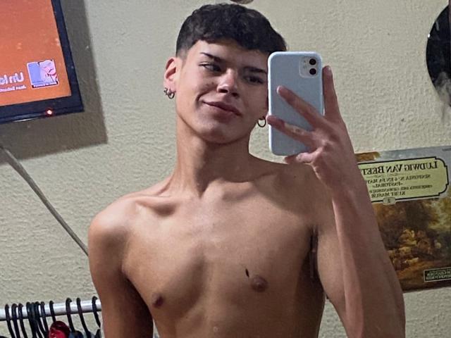 KevinMiet, shirtless, with a slim build and small nipples, holding a phone in a mirror selfie, smiling, short curly brown hair, small hoop earrings, light skin, slightly muscular.