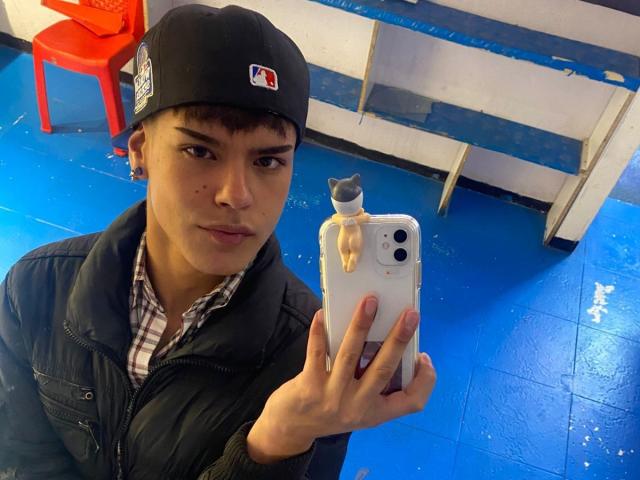 KevinMiet with a black cap, short brown hair, and a pensive face, holding a phone with a cat sticker, wearing a black jacket over a checkered shirt.