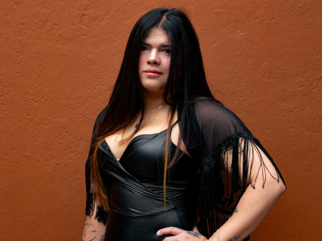 Daki Amane, com longos cabelos pretos, vestindo uma blusa preta transparente e um corpete de couro, exibindo seus seios e tatuagens, em pé em frente a uma parede laranja..