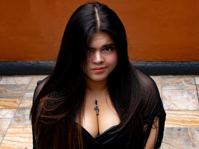 Daki Amane tiene el pelo largo y oscuro, un rostro serio y un busto grande, vestida con una blusa negra transparente, lleva un collar con un colgante, y está de pie sobre un suelo de baldosas, con una pared naranja detrás..