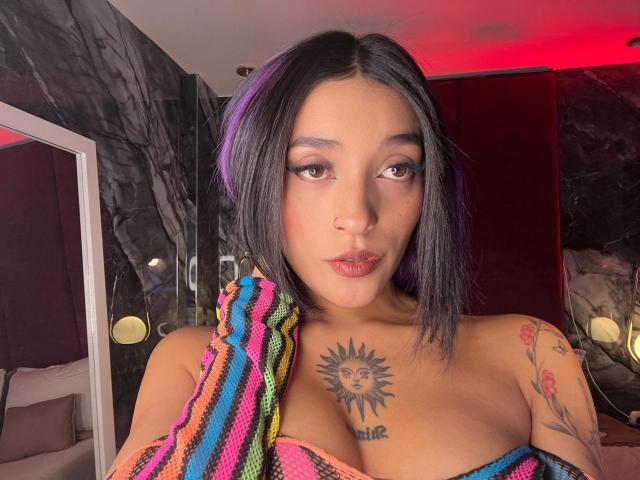 SalomeMille with black hair, medium tits, sun tattoo on chest, colorful striped top, nose ring, and tattoos, looking at camera with red lighting.