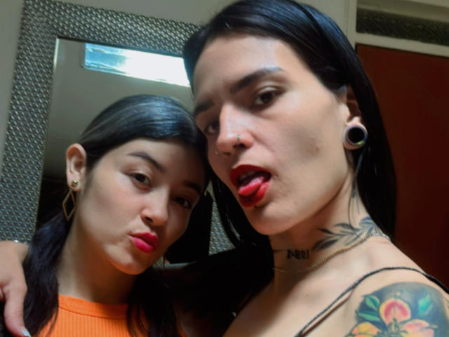 DakottaScarlett with black hair, nose ring, and tattoos, tongue out, next to DakottaScarlett in orange top, both with earrings and red lipstick, looking at mirror selfie.