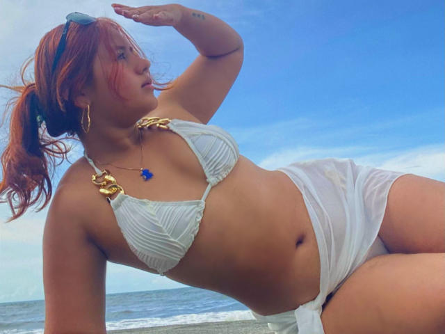 EvaLucy with red hair, wearing a white bikini top with gold straps and a white skirt, lying on a beach, blue sky, ocean in background, touching her forehead.