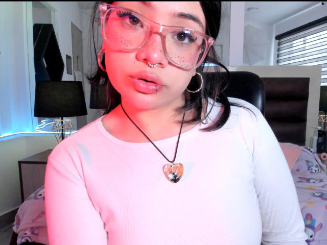 AmaraKazumi with pink glasses, nose ring, and hoop earrings, wearing a white top, heart necklace, and pink lipstick, has puffy lips and is sitting in a bedroom.