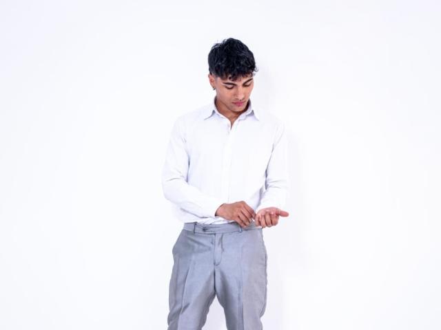 MattFiore, with short black hair, wearing a white shirt and gray pants, stands looking down, hands near his belt, against a plain white background.