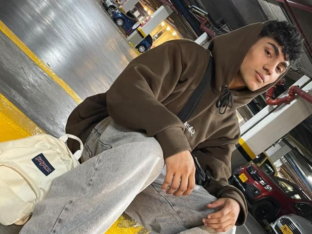 MattFiore with curly dark hair, brown hoodie, and gray pants, sitting on a yellow-painted floor, white bag next to him, red car in background, looking at camera.