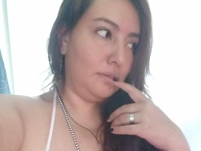 TerraMegaBoobs with brown hair, light skin, and a ring on her finger, touching her lips with a finger, wearing a silver necklace, and a white halter top.