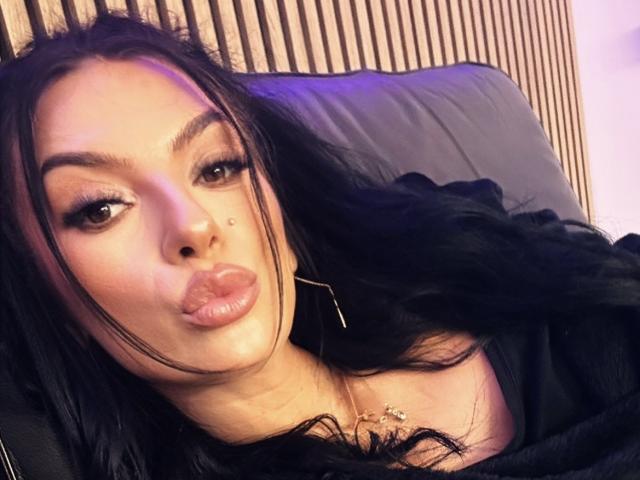 KinkyAmelie with pouty lips, black hair, and brown eyes, wearing a black top, has a small mole on her left cheek, and long earrings, looking sexy and sultry.
